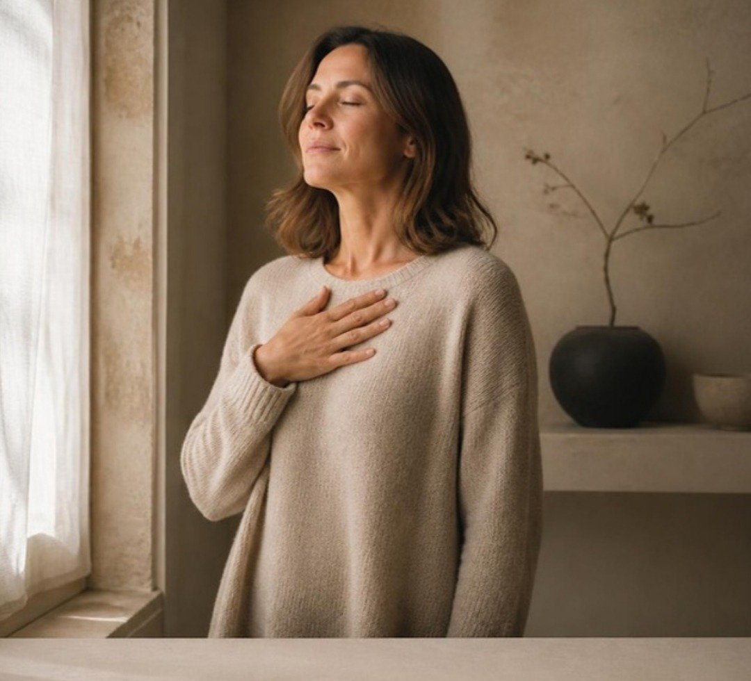 Woman in her thirties by a window, hand on her chest, in soft daylight.